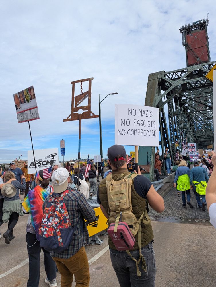 Protesters marching on a bridge at the No Kings protest in Portland, OR, one of whom is carrying a small metal guillotine on a stick.  