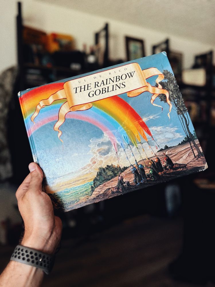 Color image. A hand holds up a well loved book. It’s Count Ul de Rico’s The Rainbow Goblins. The cover features the seven goblins lassoing a rainbow from a vibrant blue sky and forest landscape. 