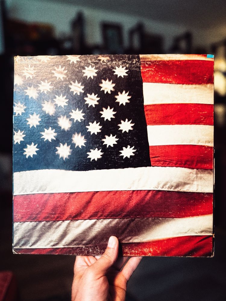 Color image. And hand holds up a battered LP sleeve. Its red white and blue colors and stars bely its musical content. It’s ’There’s a Riot Goin’ On, by Sly and the Family Stone. Released in November of 1971.