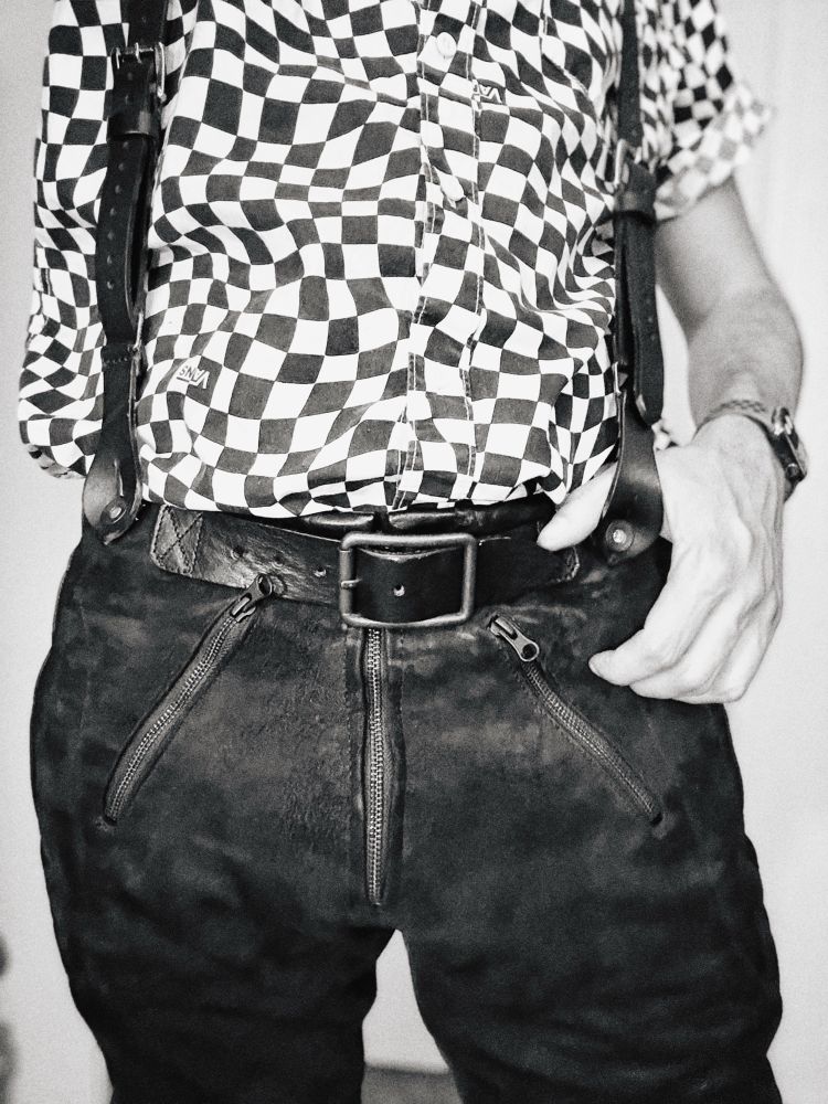 Black and white image. Low angle perspective. A tight crop on a man’s waistline. A hand rests on leather suspenders holding up his suede pants.