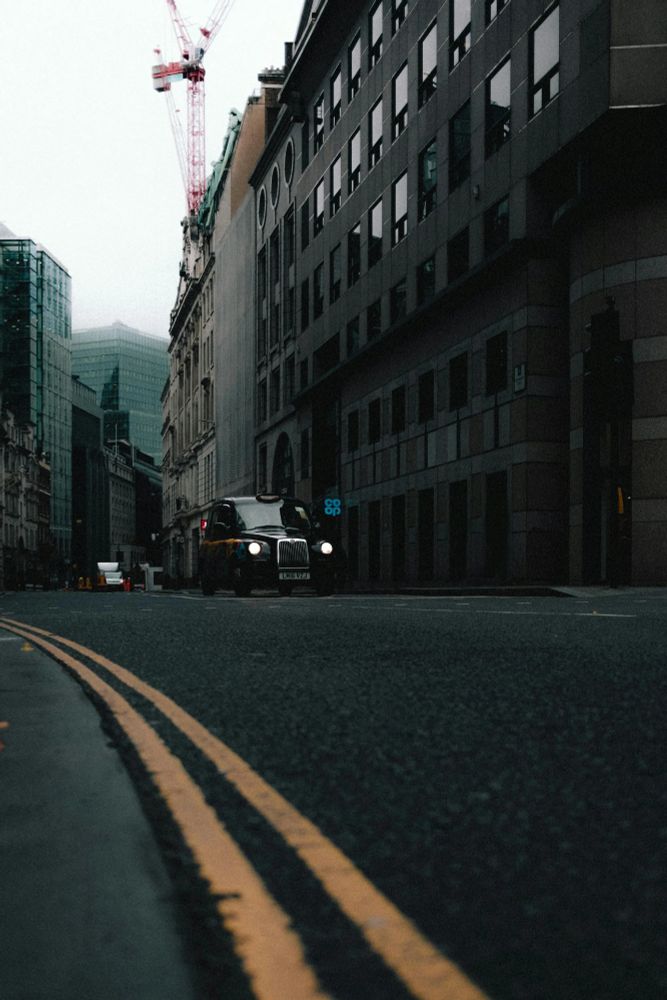 Photo by Vitor Lopes. A black taxi glides through a quiet city street, flanked by modern glass towers and older stone buildings. Overhead, a red crane looms against the pale, overcast sky, hinting at ongoing urban transformation. The double yellow lines on the asphalt draw the eye forward, emphasizing the contrast between the city's stillness and its perpetual evolution.