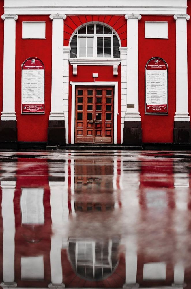 PHOTO by Artem Saranin - A striking red building with neoclassical architecture stands tall against a wet pavement, its bold facade mirrored in a shallow puddle. White columns and ornate trim frame a wooden double door beneath a half-moon window, while symmetrical posters in Russian flank each side. The reflection in the glossy surface below distorts gently, as if echoing the building’s quiet grandeur in softened ripples.