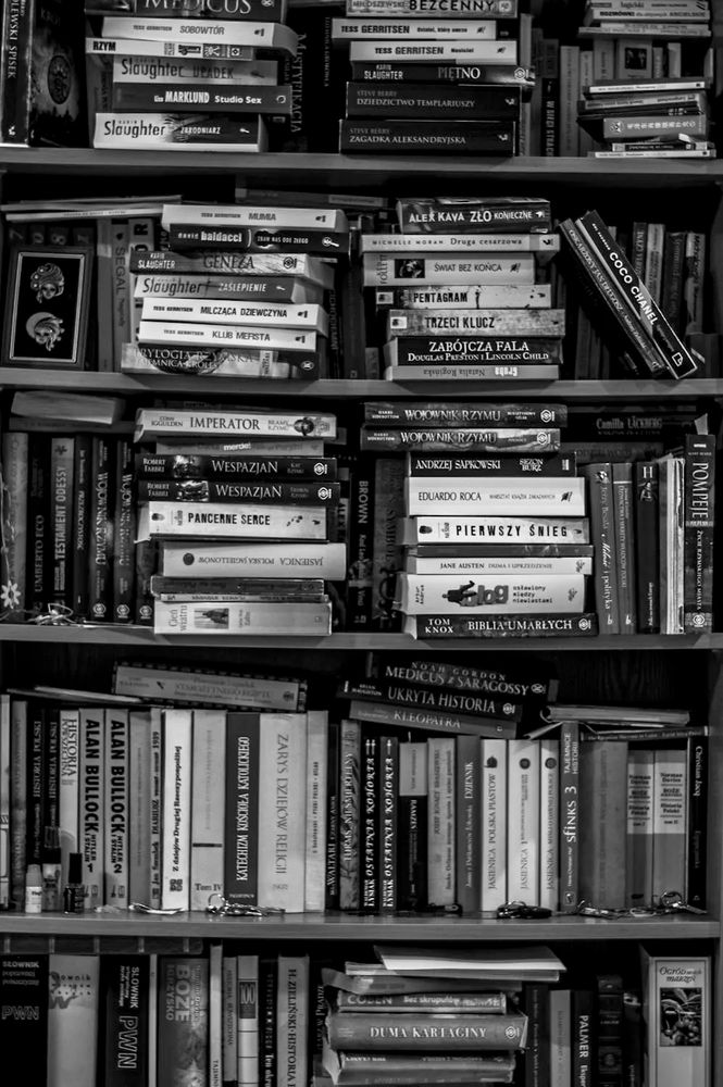 CC0 Photo. A densely packed bookshelf filled with hundreds of books arranged in various orientations—some stacked, some upright—showcasing a mix of genres, including history, thrillers, and literature. The black-and-white image highlights the texture and aged character of the well-used volumes, many of which have Polish titles, hinting at a personal or regional library.
