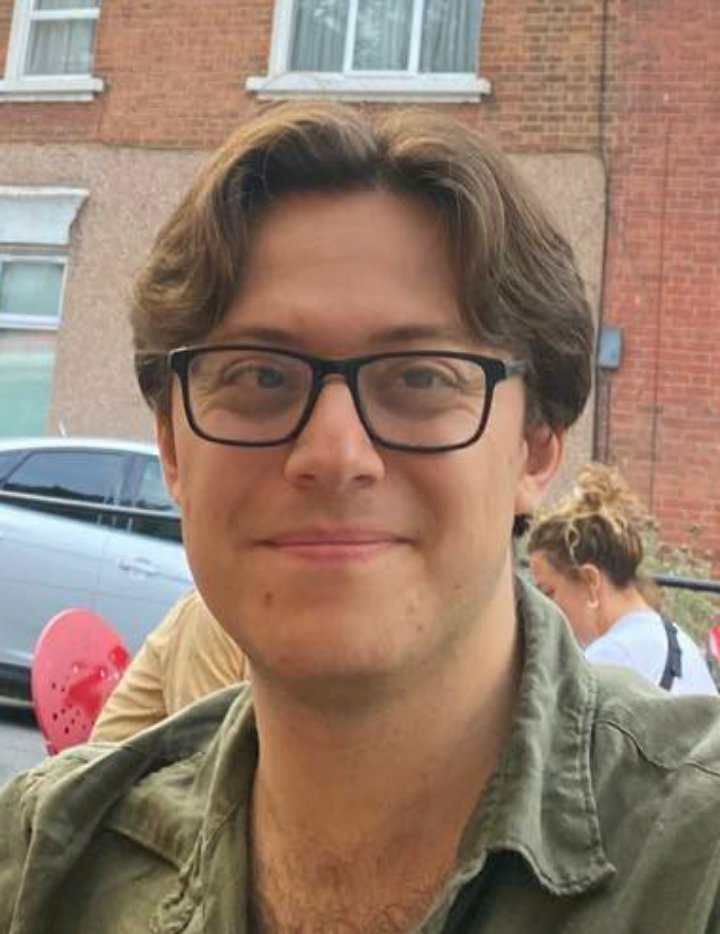 A picture of Alexander, who is looking into the camera and smiling. He is wearing glasses, a green shirt and has dark hair. There is a red brick building in the background.