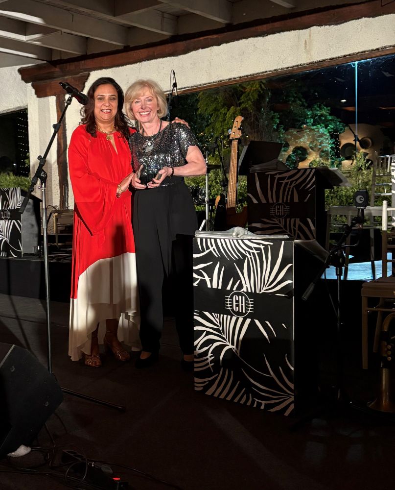 Doris is pictured receiving her award from Dipti Pandya, the Chair of EARMA. They are both standing, facing the camera and smiling. Dipti is on the left, wearing a red and white outfit. Doris is on the right wearing a black outfit.