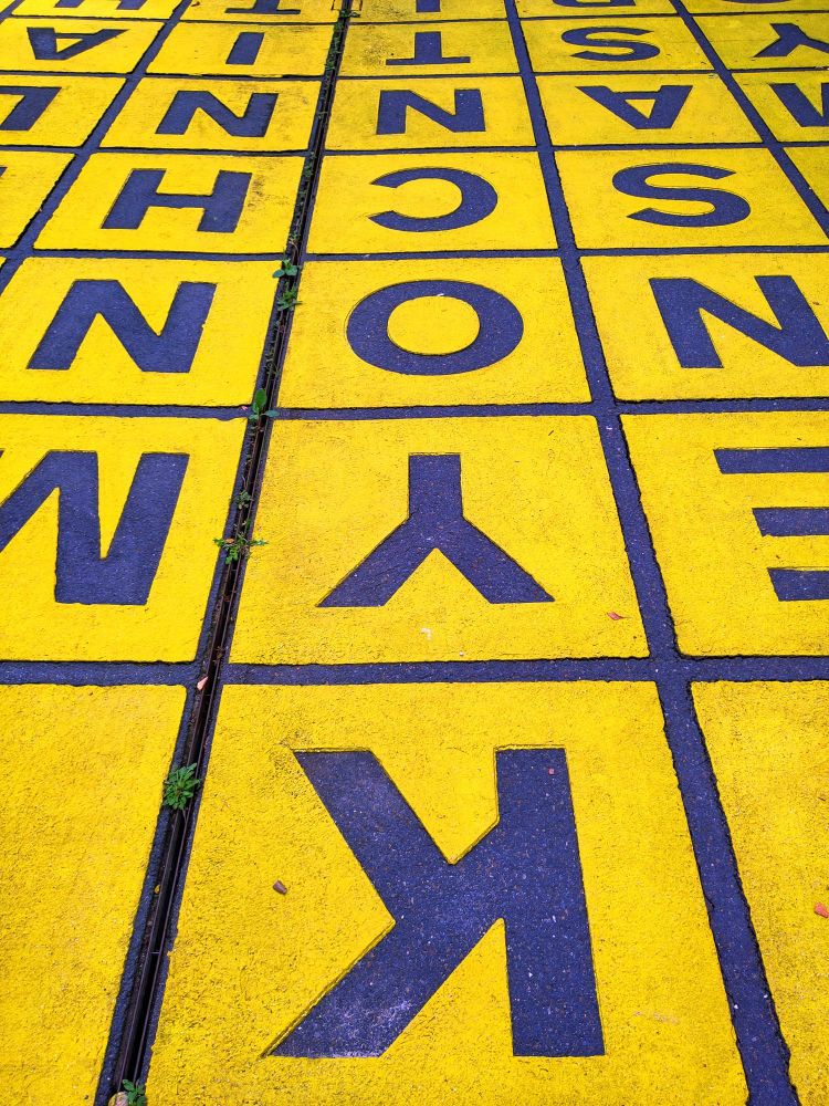 Quadratische gelbe Felder auf Asphalt mit blauen Buchstaben. Auch die quadratischen Abgrenzungen sind blau. Bildfüllend.Am Rand stehend fotografiert.