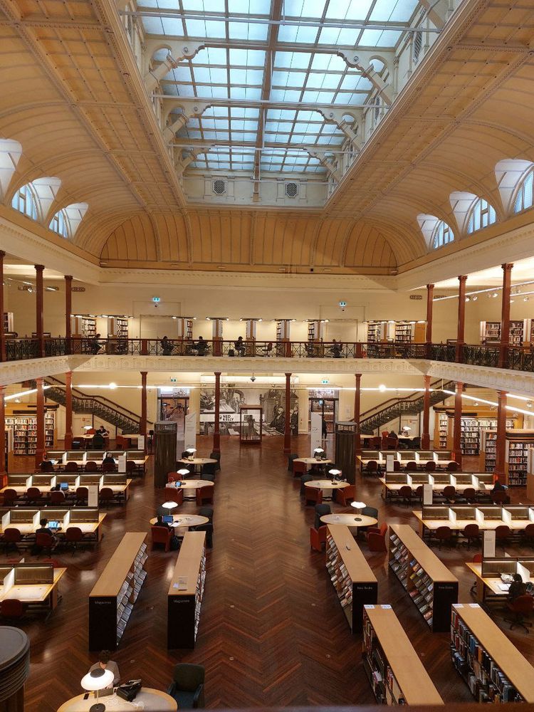 A victorian area library photographed from the second floor, showing various workplaces and bookshelves. The ceiling is made of milky glass and soft light warms the vast room.