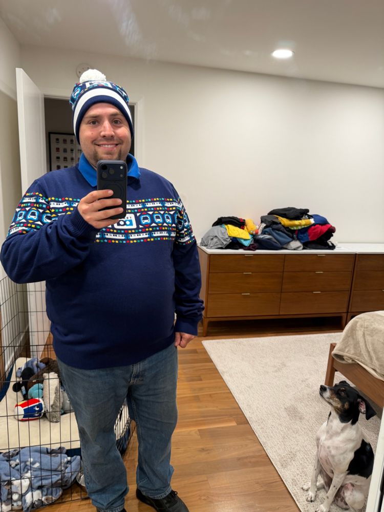 A man taking a mirror selfie while wearing a blue BART holiday sweater and beanie with white pompom. A white and black beagle mix stands to his left, looking up at him.