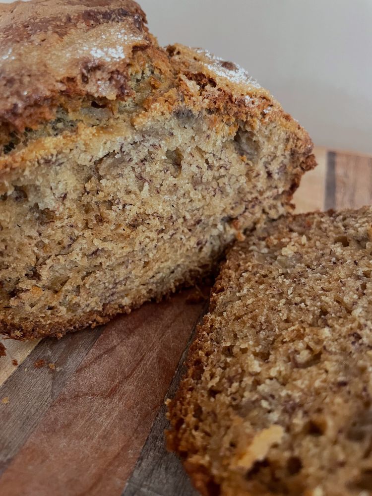 A loaf of banana bread laying on a wood cutting board with one slice cut.