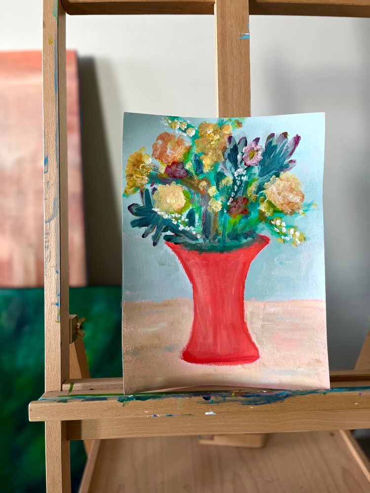 A still life painting on an easel of a bright red vase full of flowers. 