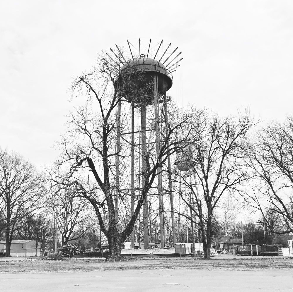 Black and white photo of a water tower with weird pipes and wires extending like a crown surrounded by a fence and trees.