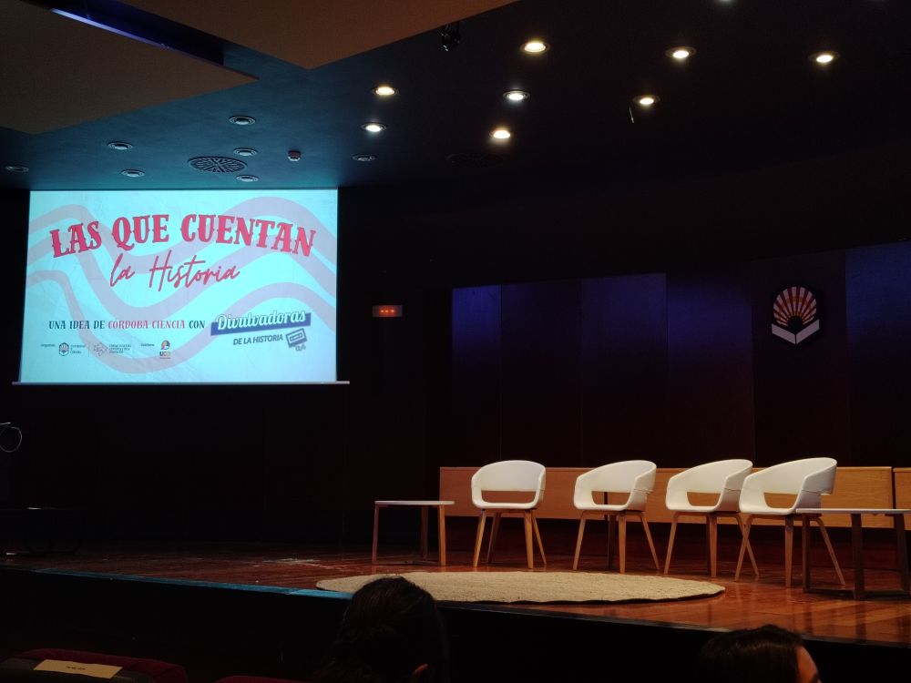 El escenario del salón de actos del Rectorado de la Universidad de Córdoba. Cuatro sillas blancas vacías en el centro, junto a una proyección del título del evento "Las que cuentan la Historia, una idea de Córdoba ciencia con Divulvadoras de la Historia". De fondo en el escenario se puede ver el emblema de la universidad de Córdoba.