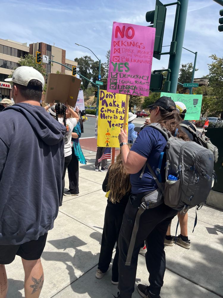 Parent and child holding homemade signs. Parent’s checklist: No Kings, Dark Money in Politics, Billionaires; Yes Democracy, Equal Rights, Liberty and Justice for All. Kid’s sign says “Don’t make me come back here in 20 years” with drawing of a child and arrows pointing to a grown up.