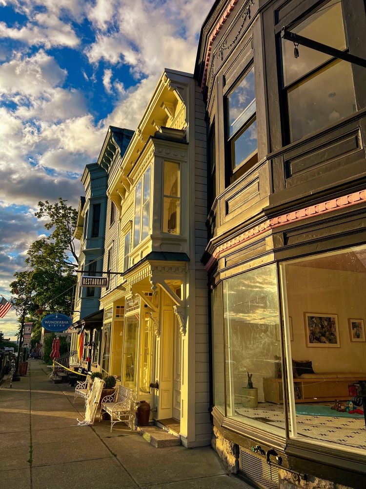 A row of beautiful old buildings in Hudson being hit straight on by the golden evening light, everything is glowing and good. Deep blue sky. Big puffy clouds with dark centers. 