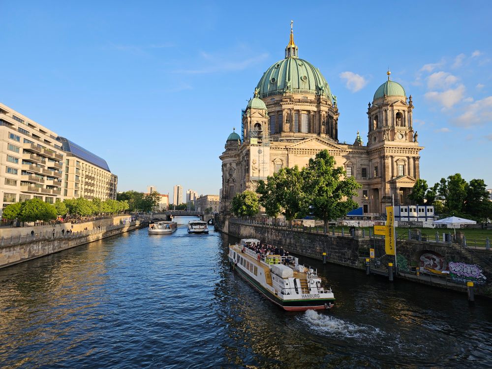Berlin cathedral on a blue sky day, the river to the left, a boat tour passing by on a nice sunny summer evening