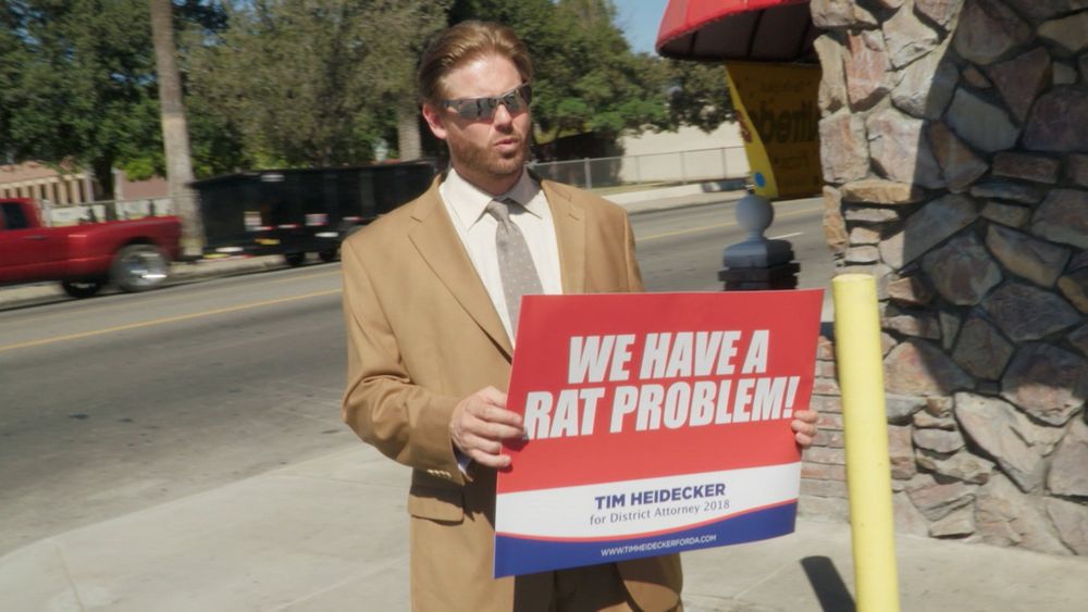 Tim Heidecker, as himself, in the movie Mister America. He’s holding a sign that says “WE HAVE A RAT PROBLEM!”