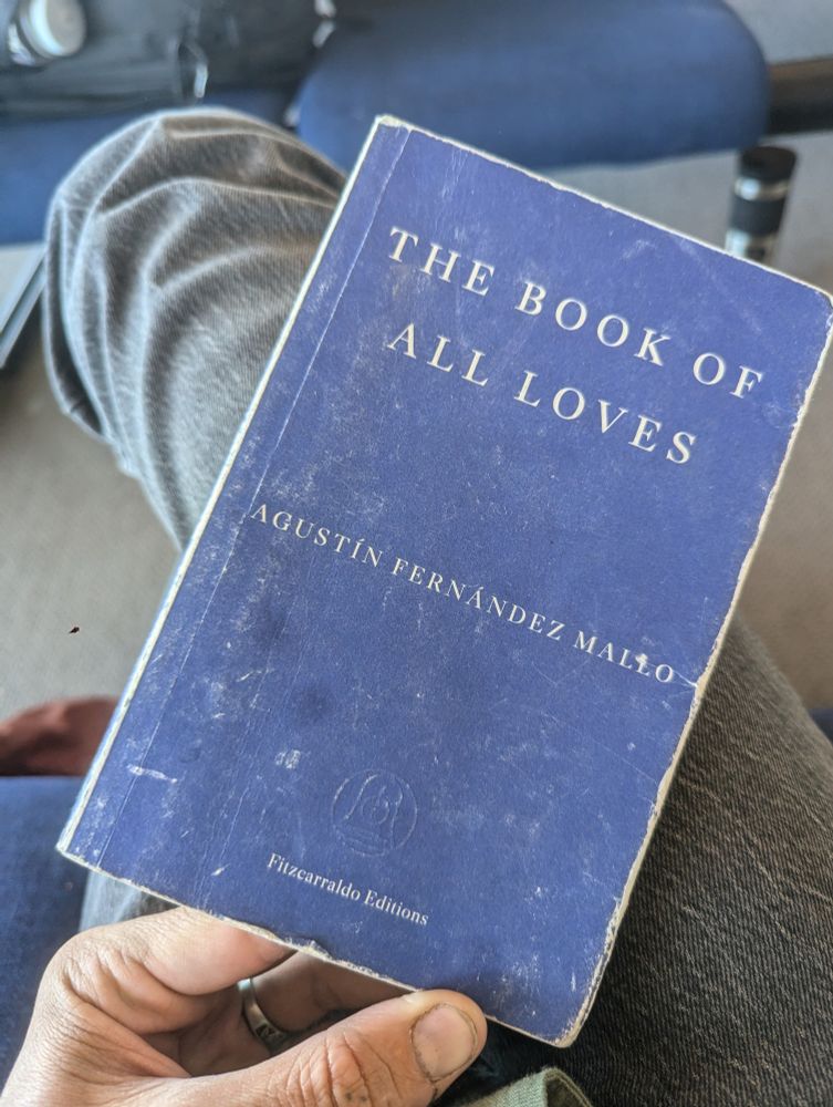 A matte blue book held on someone's knee on which the text "the book of all loves" by Agustin Fernández Mallo Is written. The book is weathered and the edges of the blue  have been torn to white. The holder is sitting on an airplane waiting lounge bench.