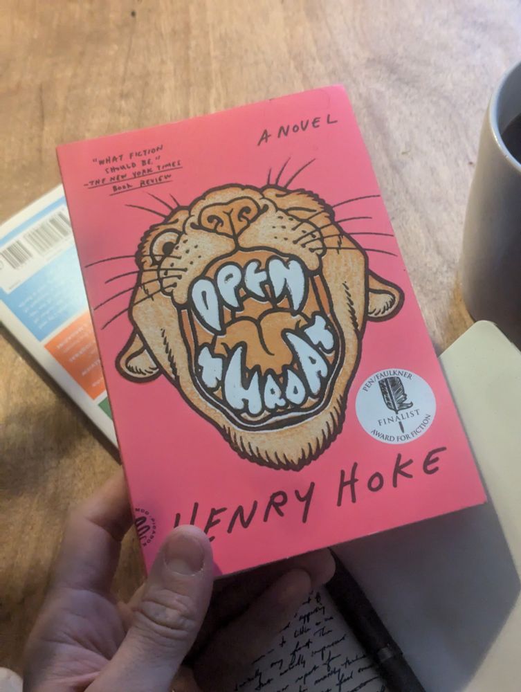 A book being held by someone's hand with the words open throat on the cover but sculpted in the form of teeth inside of a mountain lion's mouth. The book cover is Pink and the author is Henry Hoke. Underneath the book is a notebook and another book and a cup of coffee are visible in the background sitting on a wooden table.