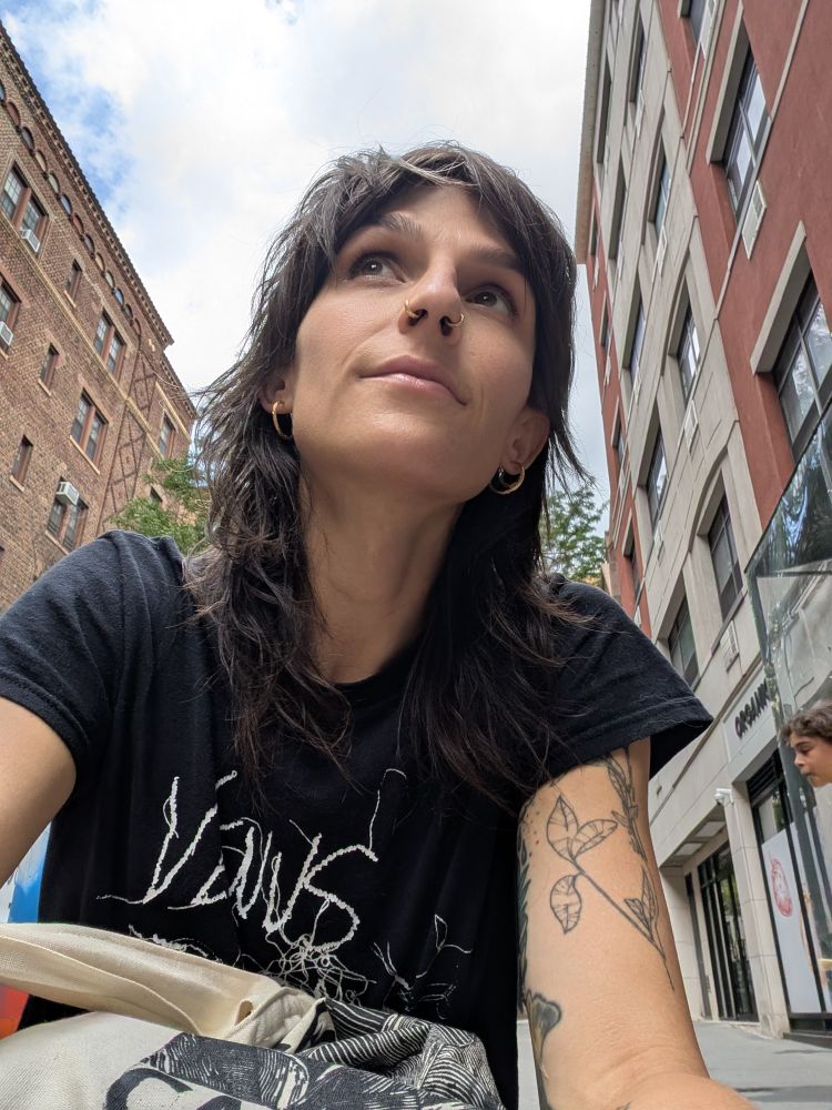 A woman wearing a black t-shirt crouches on the ground in a city setting. The photo is taken up close. She is looking above the camera. Her shirt says 'Venus Twins', from a noise punk band in Brooklyn. 