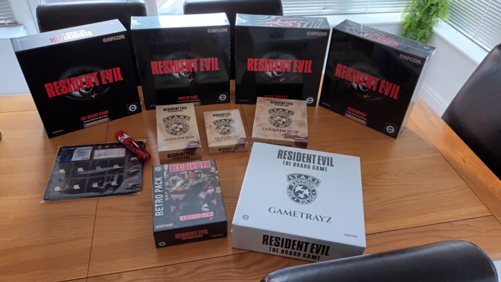 Contents of the Resident Evil Alpha Pledge Kickstarter. Base game, directors cut & 2 further expansions in black boxes with red Resident Evil text overlaid on a black and white eye. 4 additional smaller expansion packs for additional miniatures, neoprene character mats and a white box for the Gametrayz storage inserts.