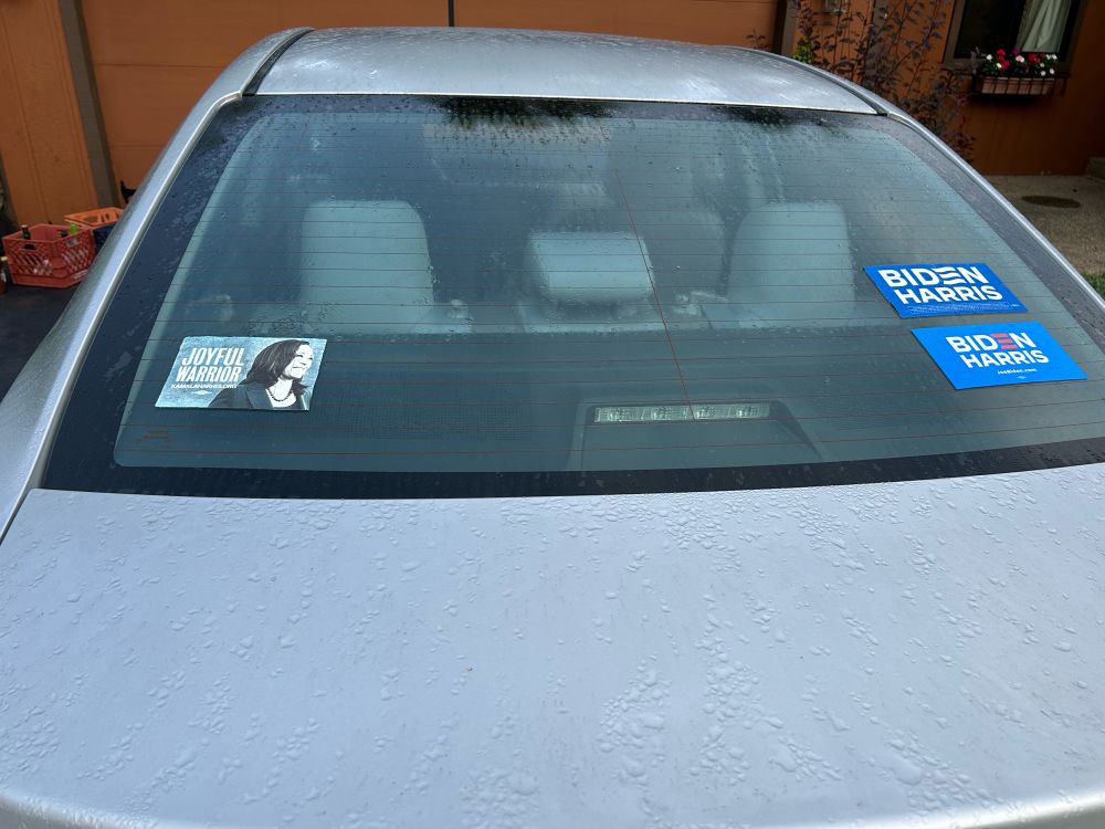 Rear window of a gray car with three political stickers:
Joyful Warrior sticker from Harris’s 2016 presidential campaign
Blue Biden/Harris from 2020
Blue Biden/Harris from 2024