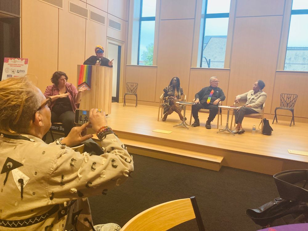 Rayceen moderates the panel from a podium. A photographer captures the scene and an ASL interpreter provided interpretation.  