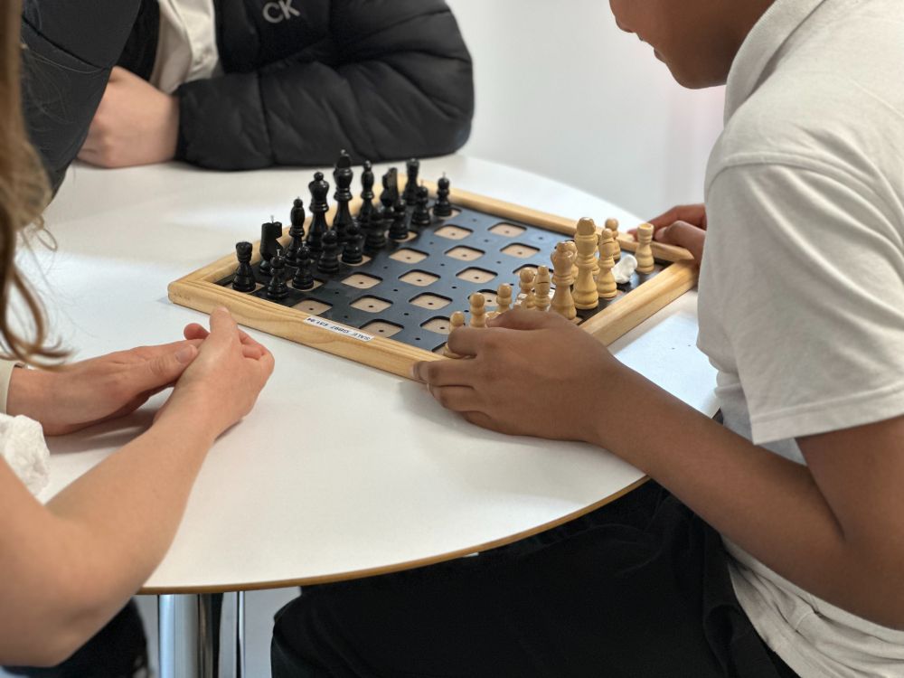 Three people sit around a round table playing a tactile chess game. The wooden chessboard has holes for peg-based pieces, making it accessible for visually impaired players. One person is moving the light-colored pieces, another observes the dark pieces, and a third pair of hands rests on the table, watching the game.
