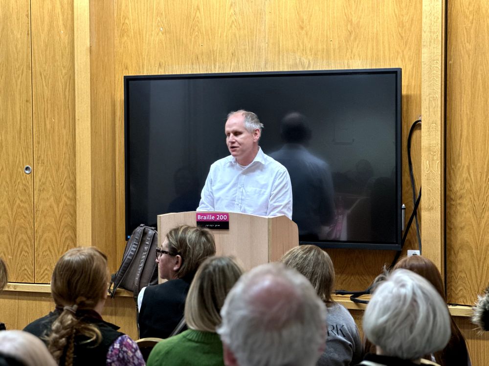 Dave from RNIB speaking at the podium in a panel room with an audience and a TV behind him, with the plaque displaying "Braille 200" in front of him.