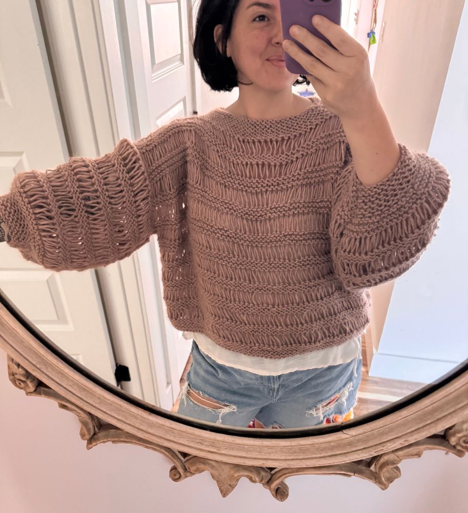 A woman stands in front of a mirror, wearing a handmade knit sweater that features long, drapey rows of stitches 