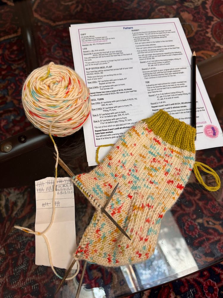 A knitted sock in progress lays on a glass table. The sock yarn is a creamy white with random flecks of hot pink, blue, green, and yellow, and the sock has a chartreuse-colored ribbing along the ankle. A printed sock pattern rests behind the knitting. 