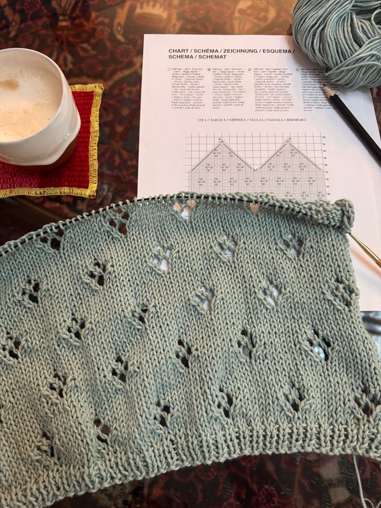 A knitting work in progress laying on a table next to a cup of coffee. The knitting features a lacy pattern with holes that form flower-like shapes. There’s a paper chart showing the knitting pattern, and a ball of light blue yarn. 