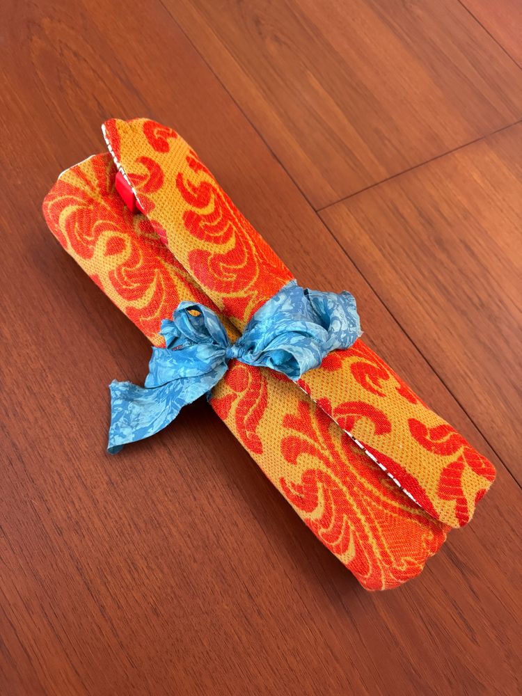 A long, rolled-up fabric case for storing knitting needles. It's made from a red and gold old-fashioned upholstery fabric, with a blue silk ribbon tied around the middle of the case to keep it closed.