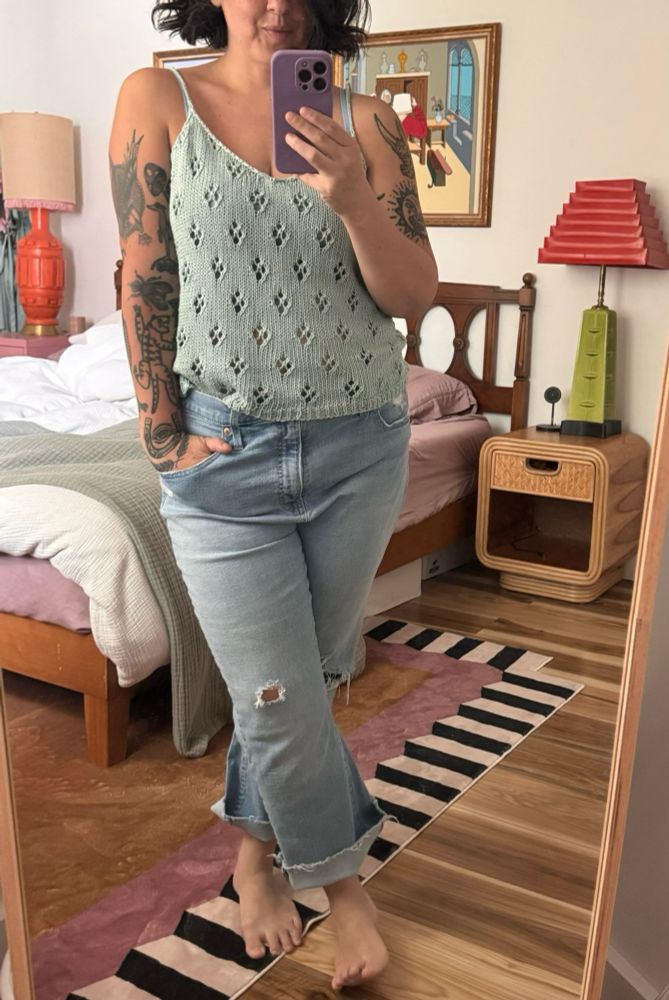 A woman stands in front of a mirror. She is wearing a handmade, light blue knit camisole. The camisole features a simple, repeated lace pattern that from a distance looks like diamond-shaped flowers. 
