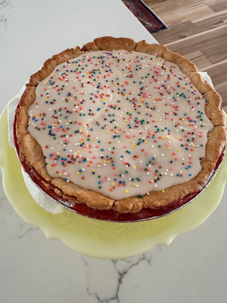 A home baked pie with a red strawberry filling and white icing with rainbow non-pareil sprinkles 