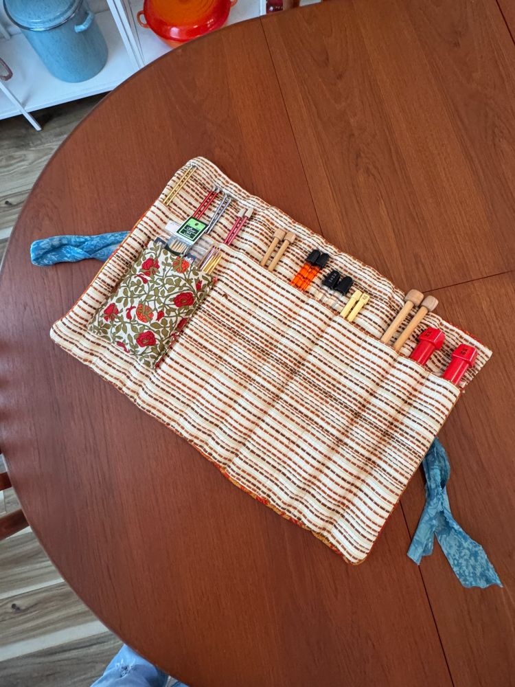A rectangular fabric case for storing knitting needles is unrolled on a table. There are pockets across the case that keep each set of needles tidy. The fabric is embroidered with red and brown stripes of thread, and features a small pocket made out of a red poppy print fabric.