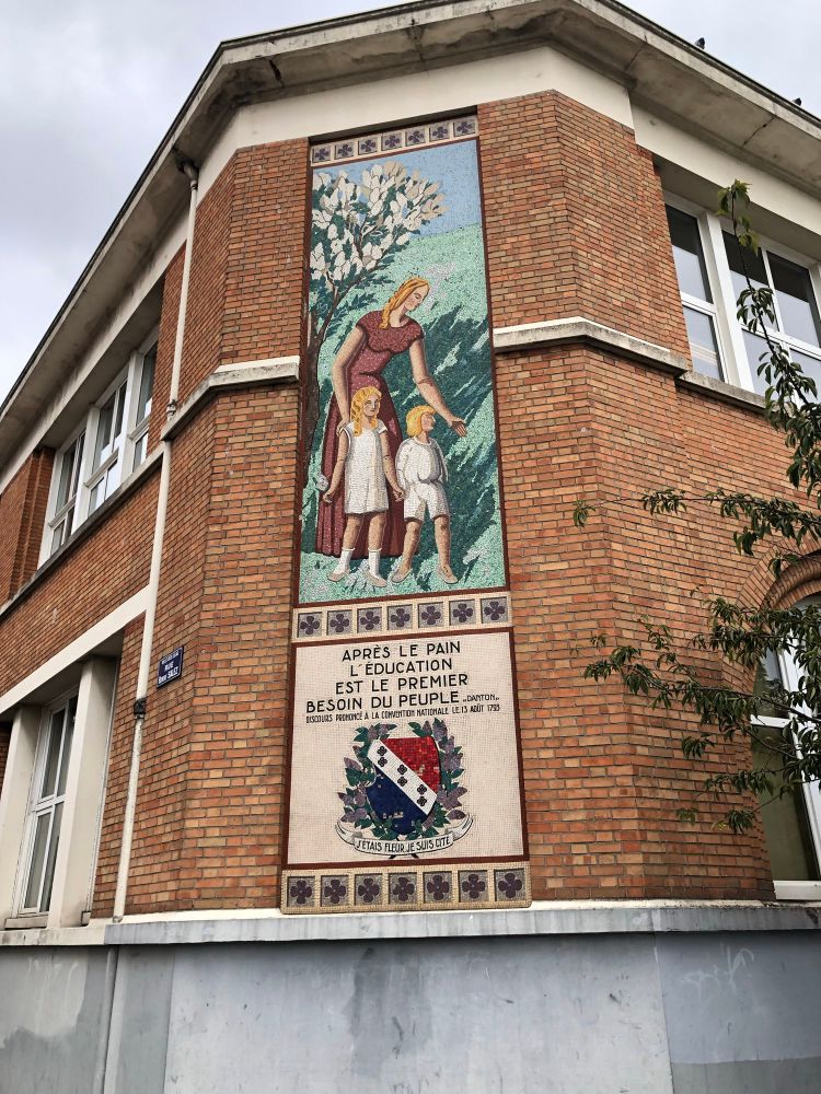 A brick elementary school building in the suburbs of Paris featuring a large vertical mosaic. The mosaic depicts a woman in a red dress guiding two children under a flowering tree. Below the scene is a quote from the French Revolution by Danton: “Après le pain, l’éducation est le premier besoin du peuple.” (“After bread, education is the first need of a people.”) The quote is accompanied by a coat of arms and the motto “J’étais fleur, je suis cité.”