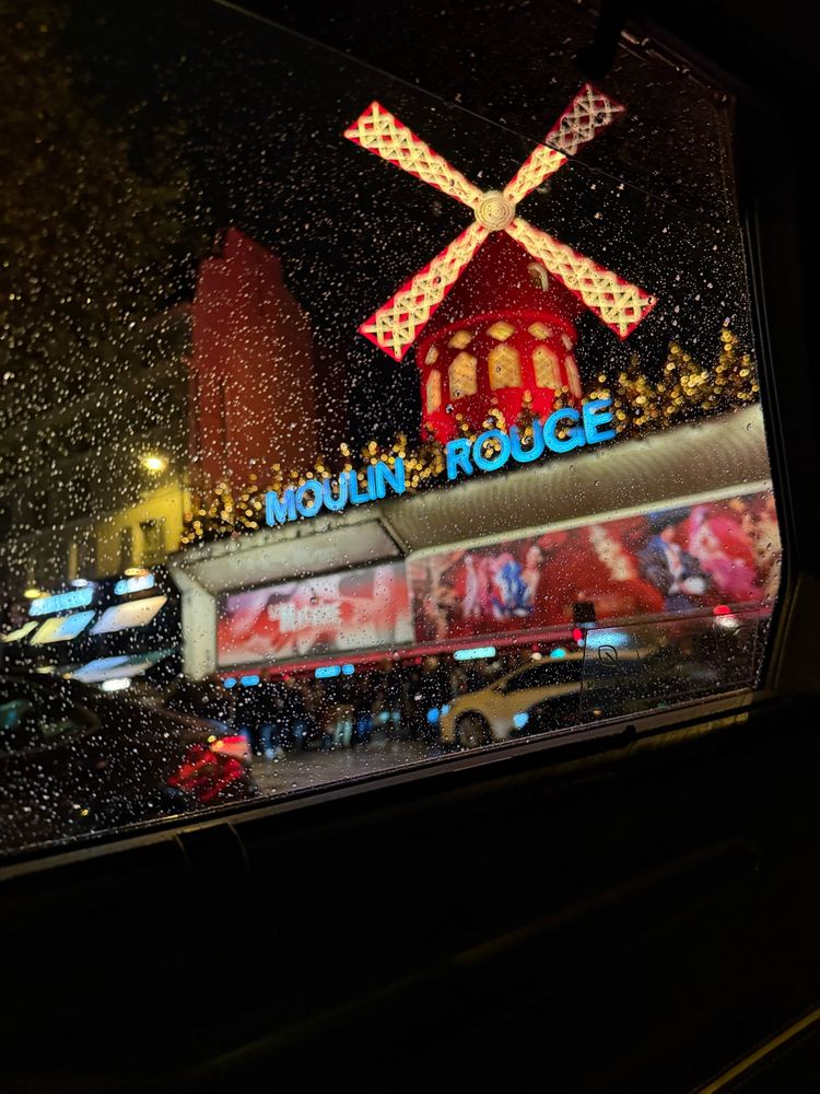 A rainy nighttime view of the Moulin Rouge, captured off-center through a cab window. The building’s iconic red windmill glows brightly against the dark, wet street.