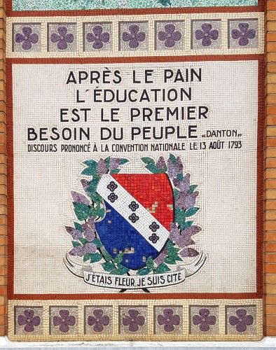 A close-up of a mosaic from a school building in the Paris suburbs. It features a quote from Georges Danton, dated August 13, 1793: “Après le pain, l’éducation est le premier besoin du peuple.” (“After bread, education is the first need of a people.”) Below the quote is a decorative coat of arms surrounded by laurel leaves and a ribbon with the phrase “J’étais fleur, je suis cité.” The border is lined with purple floral tiles.
