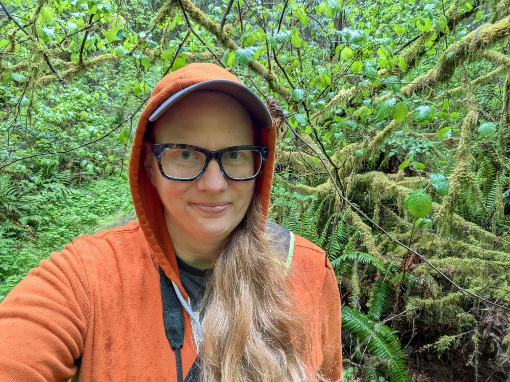 A white, blonde haired woman wearing glasses and an orange hoodie is amid a very green forest.