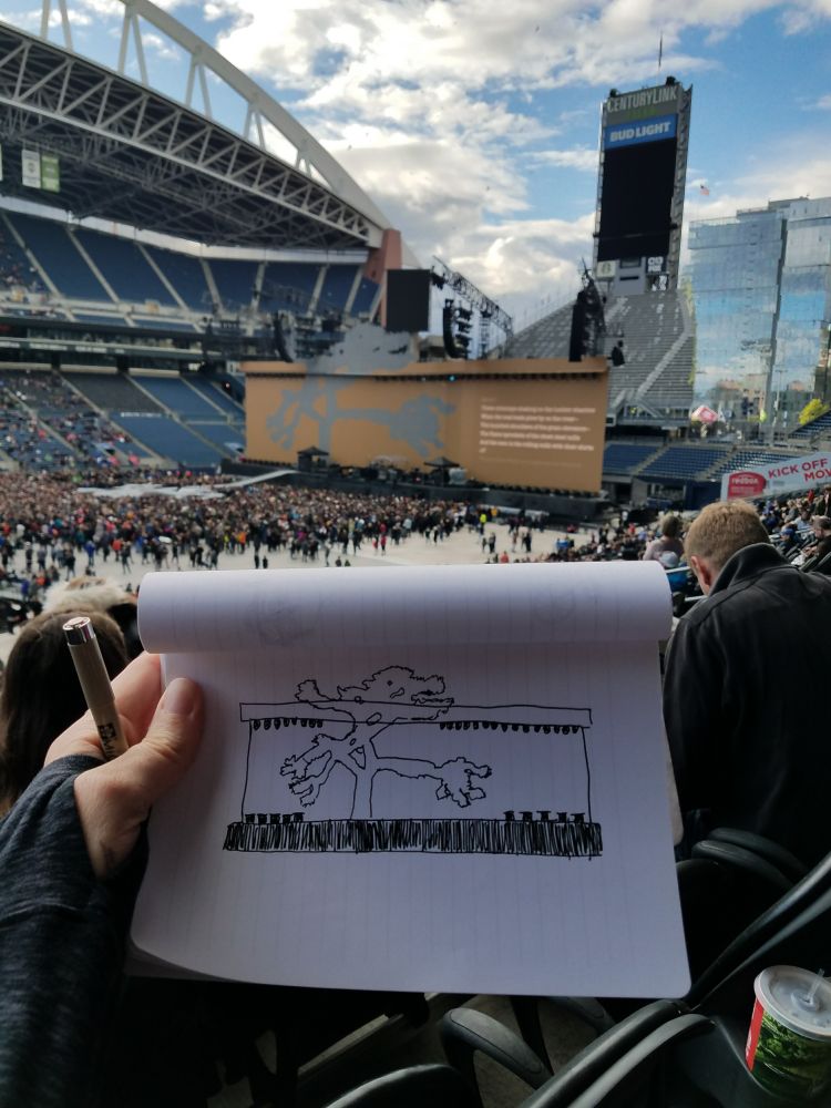 A white woman's hand holds a sketchbook and a pen. The sketchbook has a drawing of the Joshua Tree stage and is held up in front of the actual stage at Lumen Field, Seattle.