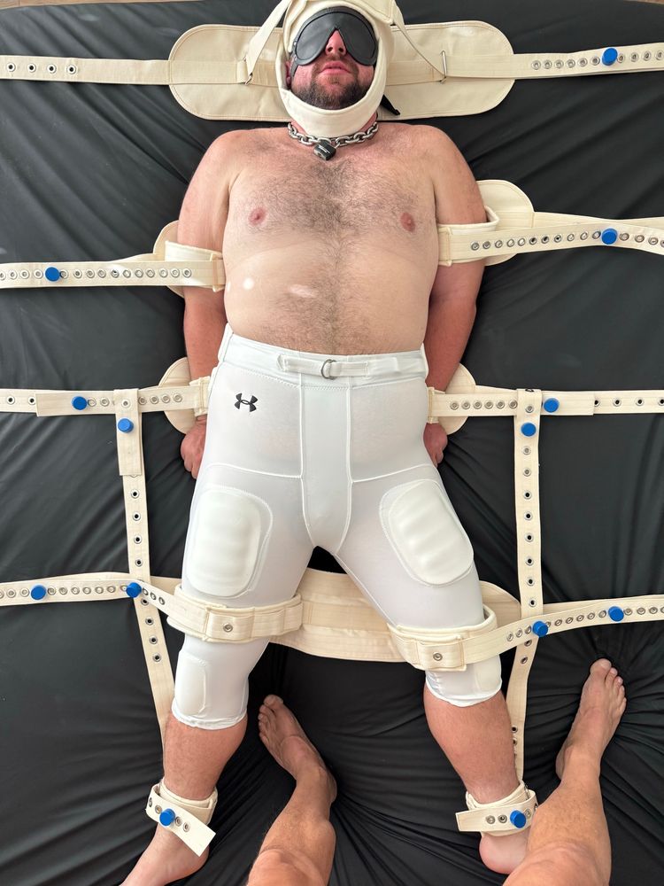 A beefy boy in white football pants is blindfolded and bound to a bed with a segufix restraint system. A man stands at his feet, looking down at his bound body.