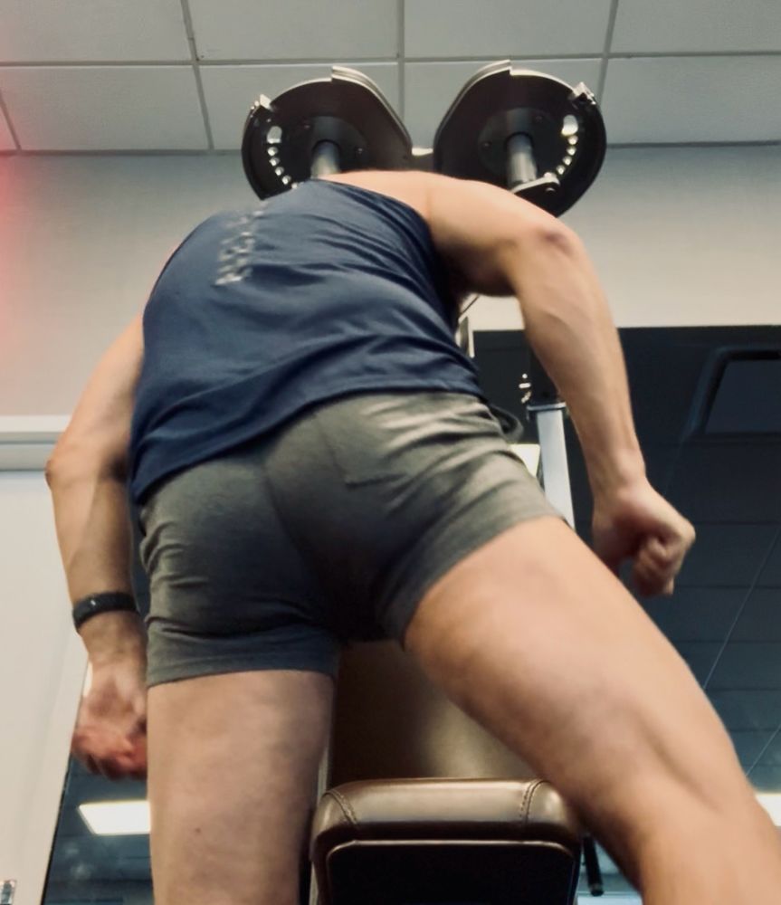 A man with a bubblebutt in tight grey gym shorts straddles a weight bench. 