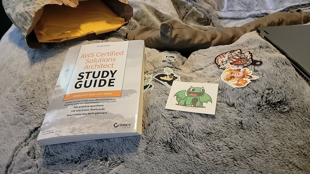 A book for studying for the AWS Solutions Architect exam, and a bunch of cute furry stickers, such as happy dragons, and a panda fursona asking for cuddles in bed