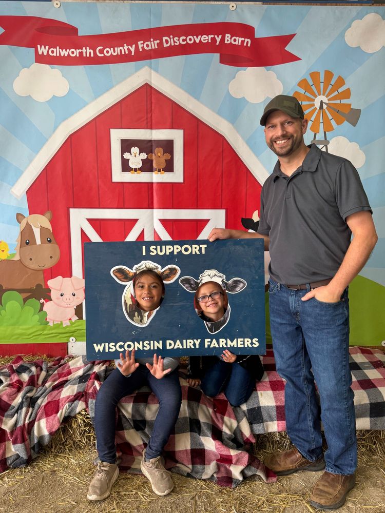 Mitchell and his daughters pose in front of a Walworth County Fair Discovery Barn backdrop. His daughters faces are posed in a cutout of cows that reads, "I support Wisconsin Dairy Farmers"