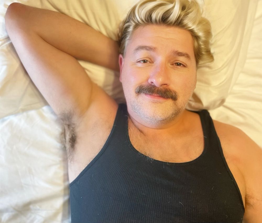 Mike Livingston posing in a black tank top and blonde wig on a bed. He also has his signature mustache.