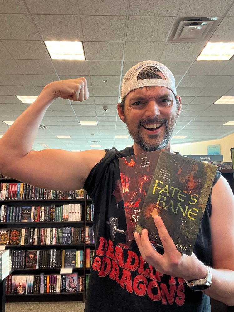 Brian posing by flexing his arm with two books, The Sovereign and Fate’s Bane, by CL Clark. He’s standing in a bookstore and wearing a shirt that says “Deadlifts & Dragons.”