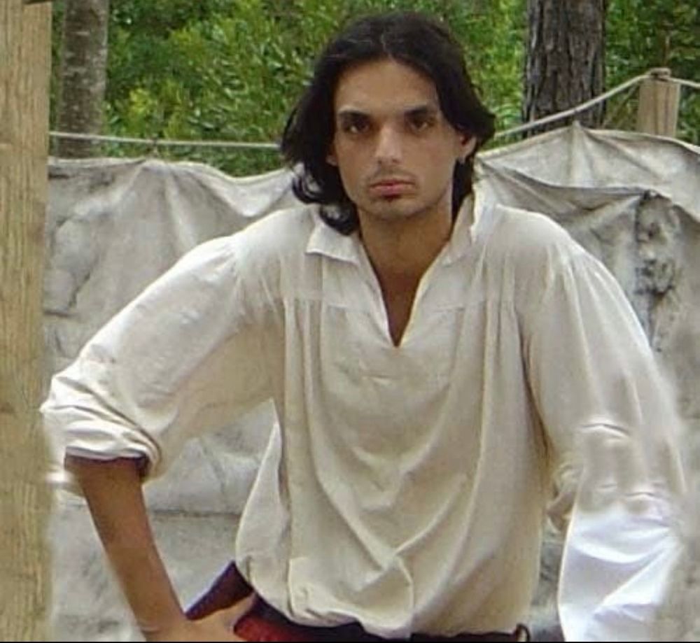 Brian in a restoration era white shirt, red linen pants. He has long black hair and stares intently at the camera. The backdrop is a tent and woods. He looks kinda like a pirate or something.