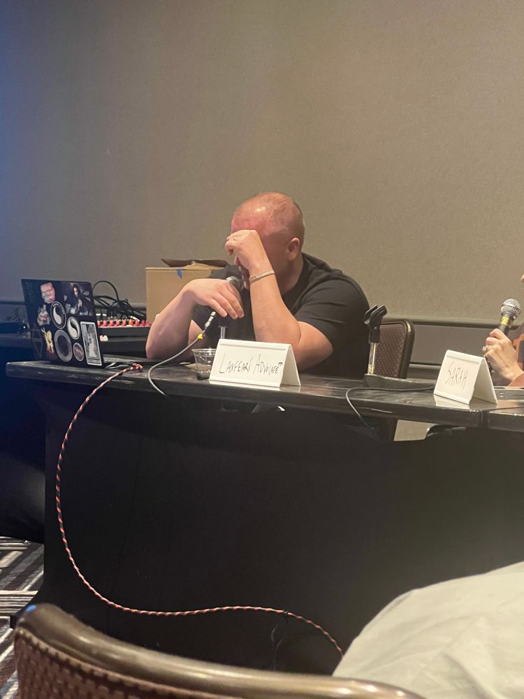 Matt sits at a table, hanging his head in embarrassment. His face is red from either laughing or crying. Either way, he’s very embarrassed