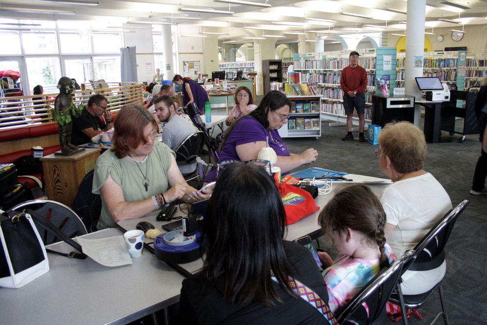 Barry library on Repair Cafe day. In the foreground are two fixers sat at a table working on different items while a child, adult and older adult wait. In the background are more tables with people having items worked on to one side and rows of books on the other.