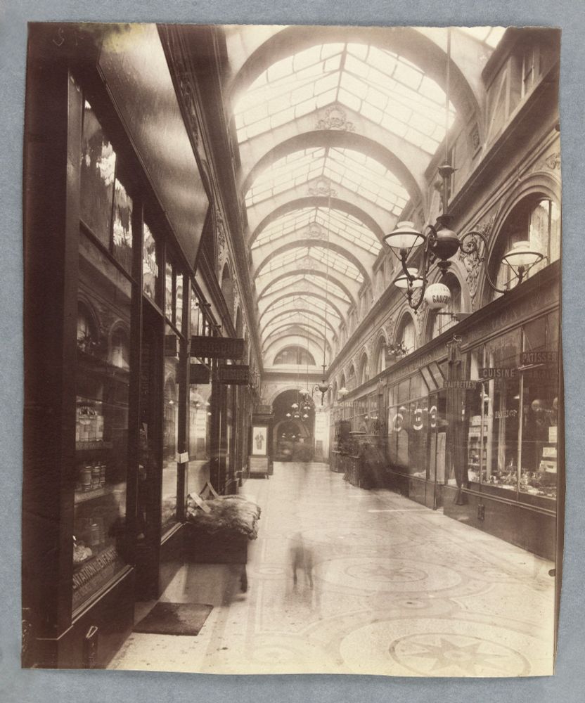 Atget's "Galerie Vivienne, 2eme ar., Paris": a black and white photograph circa 1907 of the arcades of Galerie Vivienne in the 2nd arrondissement of Paris. THe image is of a long glass covered passage with stores on either side. The image includes ghostly traces of a woman and a dog, who moved out of the image during its long exposure. 